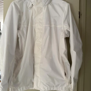 Vit vindjacka från Everest, strl 42 - Säljer en stilren vit vindjacka från Everest i storlek 42. Jackan har huva, dragkedja framtill och praktiska fickor med dragkedja. Tillverkad i lätt och tåligt syntetmaterial, perfekt för blåsiga dagar. Enkel och clean design som funkar till det mesta.Liten fläck på baksidan av jackan. 