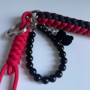 Svart pärlarmband med rosett och paracord - Stilrent armband med svarta pärlor och en söt svart rosett. Fäst i en nyckelring tillsammans med en flätad paracord-detalj i rött och svart. Perfekt accessoar att hänga på byxan eller väskan för en cool look.