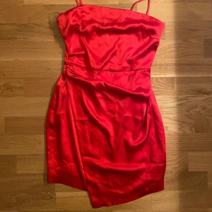 Röd kort klänning med omlott - Säljer en snygg röd kort klänning med tunna axelband och omlott-detalj i midjan. Klänningen har en figurnära passform och är tillverkad i ett glansigt material som ger en lyxig känsla. Perfekt för dig som vill sticka ut!