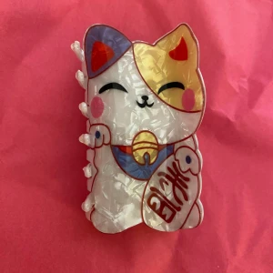 Hårklämma med Maneki Neko lyckokatt - Supergullig hårklämma i form av en Maneki Neko lyckokatt med pärlemorskimrande vit bas, detaljer i guld, blått, rött och rosa. Klämman har en tydlig kattform och japansk text på tassen. Perfekt accessoar för att piffa upp frisyren. Helt ny