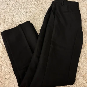 Svarta kostymbyxor från Nelly - Snygga svarta kostymbyxor från Nelly i klassisk rak modell. Byxorna har hällor för bälte, diskreta fickor bak och är tillverkade i ett mjukt tyg som känns bekvämt hela dagen. Perfekta för en clean och stilren look.