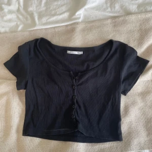 Svart croppad topp från 157 - Svart croppad topp från 157 med korta ärmar och rundad halsringning. Toppen är ribbad och har knappar framtill som snygg detalj. Perfekt för dig som gillar en enkel men trendig stil.