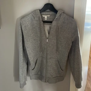 Grå hoodie med dragkedja från H&M - Mysig grå hoodie från H&M i storlek XS. Tröjan har huva med snörning, dragkedja framtill och två fickor. Perfekt för chill dagar eller när du vill ha något bekvämt. Tillverkad i mjuk bomullsblandning och har långa ärmar.