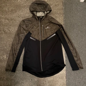 Svart och grå vindjacka från Ronhill - Snygg vindjacka från Ronhill i svart och mörkgrått med huva och hel dragkedja. Jackan har ett coolt geometriskt mönster upptill, reflekterande detaljer och en bröstficka med dragkedja. Perfekt för dig som gillar att vara aktiv utomhus.