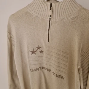 Beige stickad GANT half zip tröja - Snygg beige stickad tröja från GANT med broderad flagga och stjärnor samt texten 'GANT NEW HAVEN' på bröstet. Tröjan har hög krage, half zip-dragkedja och ribbade ärmar för en modern look.