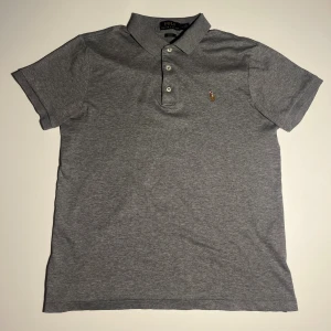 Grå pikétröja från Ralph Lauren - Klassisk grå pikétröja från Polo Ralph Lauren i slim fit-modell. Tröjan har korta ärmar, krage med tre knappar och den ikoniska broderade loggan på bröstet. Tillverkad i mjuk bomull som känns skön mot huden. Perfekt för en clean och stilren look.