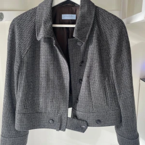 Grå skepparjacka  - Snygg grå skepparjacka från Strenesse med klassiskt rutigt mönster. Croppad modell med krage, knappar framtill och dekorativt bälte i midjan. Långärmad och tillverkad i ullblandning som ger en stilren och tidlös vibe.