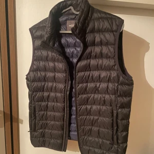 Svart dunväst från Dressmann - Stilren svart dunväst från Dressmann i storlek M. Västen har quiltad design, hög krage och dragkedja framtill. Perfekt att slänga över en hoodie eller tröja för extra värme. Lätt och smidig att bära med sig. Den är svart utanpå och mörkblå på in sidan. Det är bara att höra av dig vid frågor eller pris!