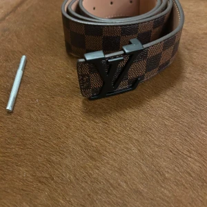 Louis Vuitton rutigt skärp i skinn - Snyggt Louis Vuitton-bälte i brunt och mörkbrunt rutigt mönster med svart LV-spänne. Bältet är gjort i skinn och har klassisk design med tydlig logga. Perfekt accessoar för att lyfta din outfit.