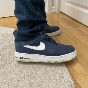 Nike Air Force 1 marinblå sneakers - Säljer ett par klassiska Nike Air Force 1 sneakers i marinblått med vit swoosh och vit sula. Skorna har snörning, perforerad tå och är tillverkade i syntetmaterial med en platt sula. Perfekta för dig som gillar streetstyle och vill ha en ikonisk look.