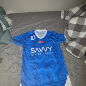 Blå Neymar Jr fotbollströja #10 - Säljer en blå fotbollströja med nummer 10 och Neymar Jr på ryggen. Tröjan har korta ärmar, vit krage och vita detaljer på sidorna. Framsidan har tryck från Savvy Games Group och Jahez, medan baksidan har Seven-logga och arabisk text. Materialet är lätt och andas, perfekt för match eller träning.