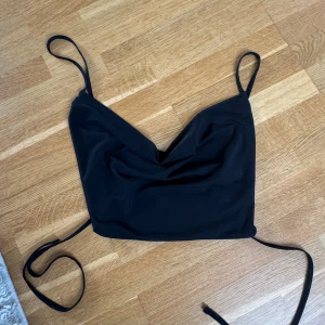 Svart croppad linne med snörning - Säljer en svart croppad linne-topp med tunna axelband och snygg snörning baktill. Toppen har en lätt draperad v-ringning och är tillverkad i ett mjukt, stretchigt material som sitter tight. 