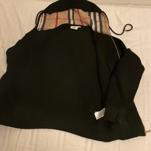 Svart hoodiejacka från Burberry M passar i S - Svart hoodiejacka från Burberry med klassiskt rutigt foder i huvan. Jackan har dragkedja i guld, ribbade muddar och en snygg, stilren look. Perfekt för dig som vill ha något både bekvämt och ikoniskt. Storlek M.