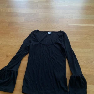 Svart ribbad topp med trumpetärm - Svart långärmad topp med ribbad struktur och vida trumpetärmar. Toppen har en rundad, lite djupare halsringning och är i mjukt, stretchigt material. 