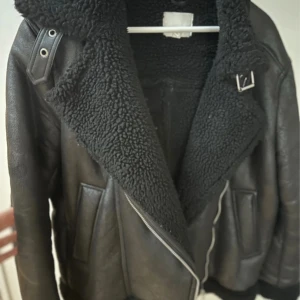 Svart teddyjacka med skinnlook - Säljer en svart teddyjacka med cool skinnlook och stora, fluffiga kragar. Jackan har dragkedja, spänne vid halsen och detaljer i metall. Insidan är fodrad med mjuk teddy och utsidan har en matt, läderliknande finish. Perfekt för vinter och passformen är oversize 