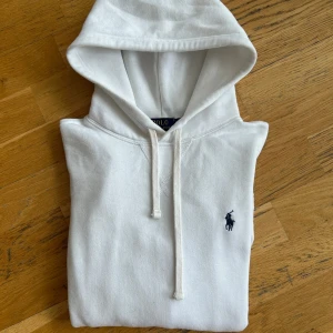 Vit hoodie från Ralph Lauren - Säljer en stilren vit hoodie från Ralph Lauren med klassisk logga på bröstet. Tröjan har en bekväm passform med känguruficka och justerbar huva med snören. 