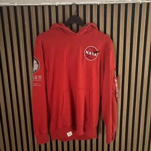 Röd Alpha Industries - Röd hoodie från Alpha Industries med NASA-logga på bröstet och Apollo 11-patch på ärmen. Har huva, känguruficka och dragkedjeficka med 'Remove Before Flight'-tag på ärmen. 