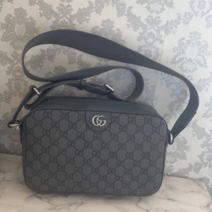 Gucci axelväska herr - Säljer en snygg axelväska från Gucci i mörkgrått skinn med klassiskt GG-mönster över hela väskan. Väskan har justerbar axelrem i textil, silverfärgade metalldetaljer och dragkedja med Gucci-logga. Perfekt storlek för det viktigaste och stilren design.