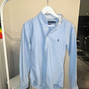Ljusblå skjorta från Ralph Lauren - Still ren ljusblå skjorta från Ralph Lauren i slim fit med klassisk krage och knappar framtill. Skjortan har lång ärm och ikoniska Polo-loggan broderad på bröstet. Tillverkad i mjuk bomull och har randigt mönster på insidan av kragen för extra detalj. PRIS KAN DISKUTERAS 