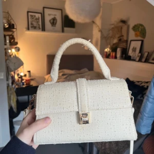 Beige handväska i bomullstyg - Säljer en stilren beige handväska i bomullstyg med struktur. Väskan har ett topphandtag, avtagbar axelrem och snyggt guldfärgat lås framtill. Perfekt storlek för det viktigaste och enkel att matcha med olika outfits. 💕