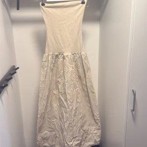 Lång beige axelbandslös klänning - Säljer en stilren långklänning i beige med axelbandslös design. Klänningen har en figurnära överdel och en vid, rynkad kjol som ger en luftig känsla. Perfekt för sommarens varma dagar eller speciella tillfällen. Enkel och tidlös modell som är lätt att matcha.