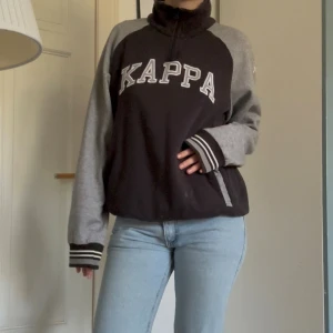 Svart och grå tjocktröja med krage från Kappa - Säljer en svart och grå tjocktröja från Kappa med broderad logga framtill. Jackan har långärmade grå ärmar, ribbade muddar med ränder och halv dragkedja vid kragen. Perfekt för dig som gillar sportig och avslappnad stil.
