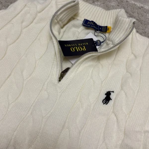 Vit kabelstickad tröja Polo Ralph Lauren - Kabelstickad vit tröja från Polo Ralph Lauren med halv dragkedja och broderad logga på bröstet. Tröjan har lång ärm, ribbade muddar och hög krage. Perfekt för lager-på-lager och snygg till jeans eller kjol.