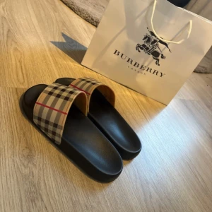 Burberry sandaler med klassiskt rutmönster - Snygga sandaler från Burberry med den ikoniska beigea och svarta rutan samt röda detaljer. Svart platt sula med Burberry-logga på sidan. Perfekta för sommaren och riktigt trendiga att glida runt i. Tillverkade i syntetmaterial.