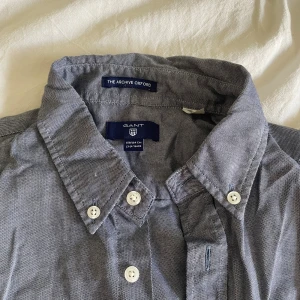 Grå oxfordskjorta från Gant - Snygg grå oxfordskjorta från Gant med klassisk button down-krage och vita knappar. Skjortan har lång ärm och bröstficka med broderad Gant-logga. Perfekt för dig som gillar stilrena och tidlösa plagg.