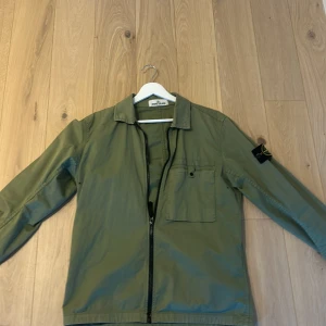 Grön overshirt från Stone Island - Snygg militärgrön overshirt från Stone Island med klassisk patch på ärmen. Jackan har dragkedja framtill, bröstficka med knapp och krage. Perfekt för lager-på-lager. Tveka inte på att höra av er för flera bilder!