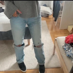 Ljusblå slitna jeans med hål på knäna - Säljer ett par ljusblå jeans med raka ben och stora hål över båda knäna. Jeansen har klassisk femficksdesign och är tillverkade i denim. Perfekta för en avslappnad och trendig look.