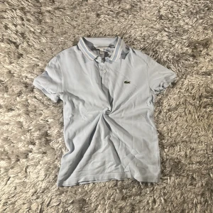 Ljusblå pikétröja från Lacoste - Ljusblå pikétröja från Lacoste med klassisk krage och knappar framtill. Slim fit-modell med den ikoniska gröna krokodilloggan broderad på bröstet. Tröjan är tillverkad i bomull och har korta ärmar samt diskreta vita ränder på kragen.