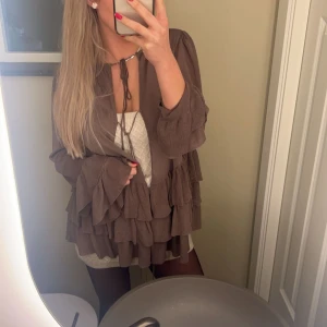 Tröja med volanger  - Brun tröja med volanger i lite syntetiskt material. Älskar älskar denna tröjan men råkade krympa lite i tvätten: den var en M men är nu en liten S/XS. Kan knytas i 3 snörningar på framsidan som man ser på bilden 🌟 vill man ha lite större passform så passar den en XS.