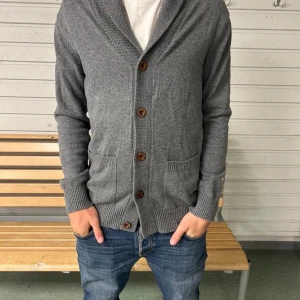 Grå kofta perfekt till höst/ vintern  - Mysig grå kofta från Jack & Jones med sjalkrage, bruna knappar och två fickor fram. Snygga bruna mockadetaljer på armbågarna. Tillverkad i bomull, akryl och ull – perfekt för lager på lager under kyliga dagar.