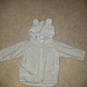 Teddyjacka med öron - Supersöt vit teddyjacka för nyfödd med huva och små öron på toppen. Jackan är fluffig och mjuk, har dragkedja framtill och är helfodrad med ett diskret mönster på insidan. Perfekt för att hålla din lilla varm och gullig under kyliga dagar. Finns en fläck framtill som jag provat att få bort!