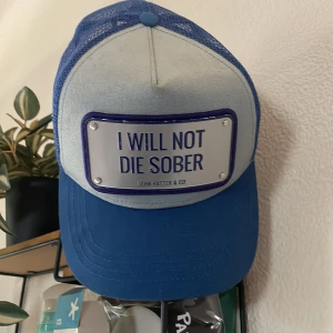 Blå truckerkeps John Hatter & Co - Snygg blå truckerkeps från John Hatter & Co med vit frontpanel och blått mesh-tyg baktill. Framsidan har en metallskylt med texten 'I WILL NOT DIE SOBER'. Justerbar snapback och klassisk böjd skärm. Perfekt statement-piece för dig som vill sticka ut.