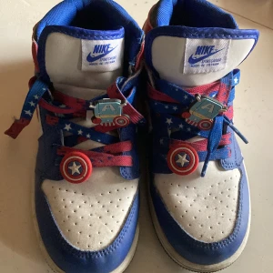 Nike Air Jordan 1 Captain America sneakers - Nike Air Jordan 1 sneakers i Captain America-tema med blå, vita och röda paneler i skinn. Snygga snören med stjärnmönster och coola Captain America-dekorationer. Klassisk hög modell med rund tå och platt sula. Perfekt för dig som vill sticka ut med Marvel-vibe.