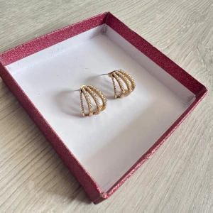 Guldiga örhängen med pärldetaljer - Unika örhängen i guldigfärg med små vita pärlor längs varje båge. Aldrig använda och i topp skick 