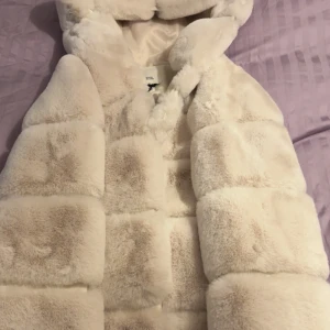 Beige pälsjacka från River Island - Fluffig och lyxig beige pälsjacka från River Island med stor huva och quiltad look. Västen har ett mjukt, glansigt innerfoder och breda paneler som ger en cool och trendig vibe. Perfekt för att lyfta din outfit under kyliga dagar. Jackan är helt ny aldrig använd: Nypris 1000kr