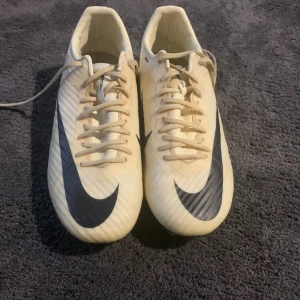 Nike Mercurial vita fotbollsskor - Säljer ett par Nike Mercurial fotbollsskor i vit färg med svarta detaljer och stora swoosh-loggor. Skorna har snörning och platt sula med fasta dobbar för gräsplan. Lätta och smidiga med syntetmaterial för bästa bollkontroll. Perfekta för dig som vill sticka ut på planen.
