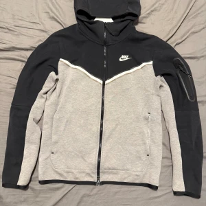 Nike tech fleece svart och grå  (S) - Nike hoodie i svart och grått med hel dragkedja och huva. Fleecen har en vit Nike-logga på bröstet, praktiska fickor och en extra ficka med dragkedja på ärmen. Tillverkad i mjuk bomullsblandning, perfekt för en sportig och avslappnad stil. Nypris 1200kr. Pris kan diskuteras😊