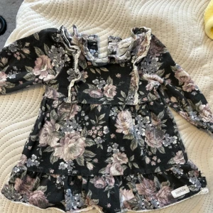 Blommig volangklänning från Newbie - Supersöt kortklänning från Newbie med mörk botten och blommigt mönster i lila, rosa och beige toner. Klänningen har volangdetaljer vid axlar och nederkant, långärmade ärmar med resår och spetskant vid halsen.