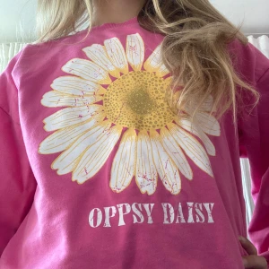 Rosa sweatshirt med stor prästkrage - Superfin rosa sweatshirt från Cubus med stor prästkrage-tryck i vitt och gult samt texten 'OPPSY DAISY' framtill. Tröjan har rund hals, långa ärmar och ribbade muddar. Perfekt för dig som gillar färg och blommiga prints! 💖✨✨🫶🏻🥰💞