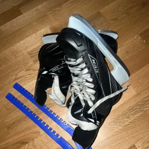 Svarta CCM skridskor med snörning - Säljer ett par svarta CCM skridskor med vita detaljer och grå snören. Skridskorna har en robust design med vit sula och klassisk snörning. Perfekta för isen och kommer med blå skydd till skenorna. Snygg och sportig look för vintern!