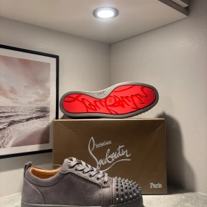 Grå Christian Louboutin sneakers med spikar - Säljer ett par gråa Christian Louboutin sneakers i mocka med ikonisk röd sula och coola nitar framtill. Skorna har snörning och rund tå, samt exklusiv känsla med lyxiga detaljer. Perfekta för dig som vill sticka ut med stil. Hör av vid fundering