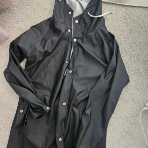 Marinblå regnjacka från 157 med huva - Marinblå regnjacka från 157 i storlek XS. Jackan har huva med vit snörning, tryckknappar framtill och två sidofickor. Materialet är syntet och jackan är perfekt för blöta dagar. Enkel och stilren design med längre passform.