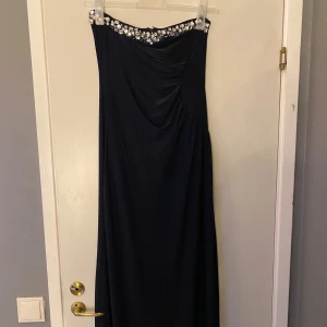 Marinblå långklänning med strass - Snygg marinblå långklänning med axelbandslös design och glittrande strassdetaljer längs överdelen. Klänningen har draperade partier framtill och en diskret dragkedja baktill. Materialet är mjukt och har en lätt glansig finish som ger en lyxig ❤️känsla. Frak ingår inte 