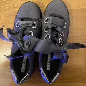 Grå coola sneakers  -   I storlek 36. Snygga grå sneakers från Malien med breda satinliknande snören och strass runt öljetterna. Skorna har rund tå, platt sula och en metallic-detalj på hälen. Perfekta för dig som vill ha något extra till din outfit.
