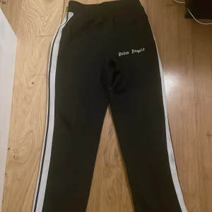 Palm Angels svarta trackpants med vita ränder - Svarta trackpants från Palm Angels med vita ränder längs sidorna och logga tryckt på framsidan. Byxorna har elastisk midja och är tillverkade i polyester för en sportig vibe. Perfekta för en streetwear-look.