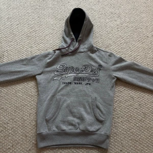 Superdry hoodie - Säljer nu en ricktigt snygg grå hoodie från Superdry. Det finns ett logotyptryck på bröstet. Tröjan har huva med snöre. Skicket är som nytt så skulle säga 9/10. Det är bara att höra av er vid frågor🤩🤩